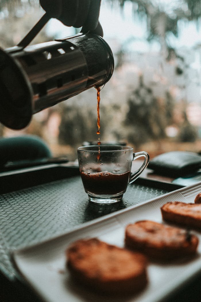 Hakkımzda Aromatic coffee being poured into a glass cup beside a plate of biscotti on a tray.